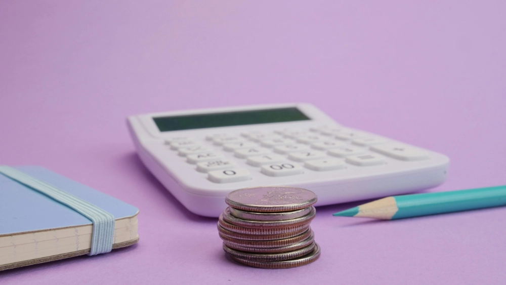 Picture of a calculator, blue pencil and a stack of change with a purple background.