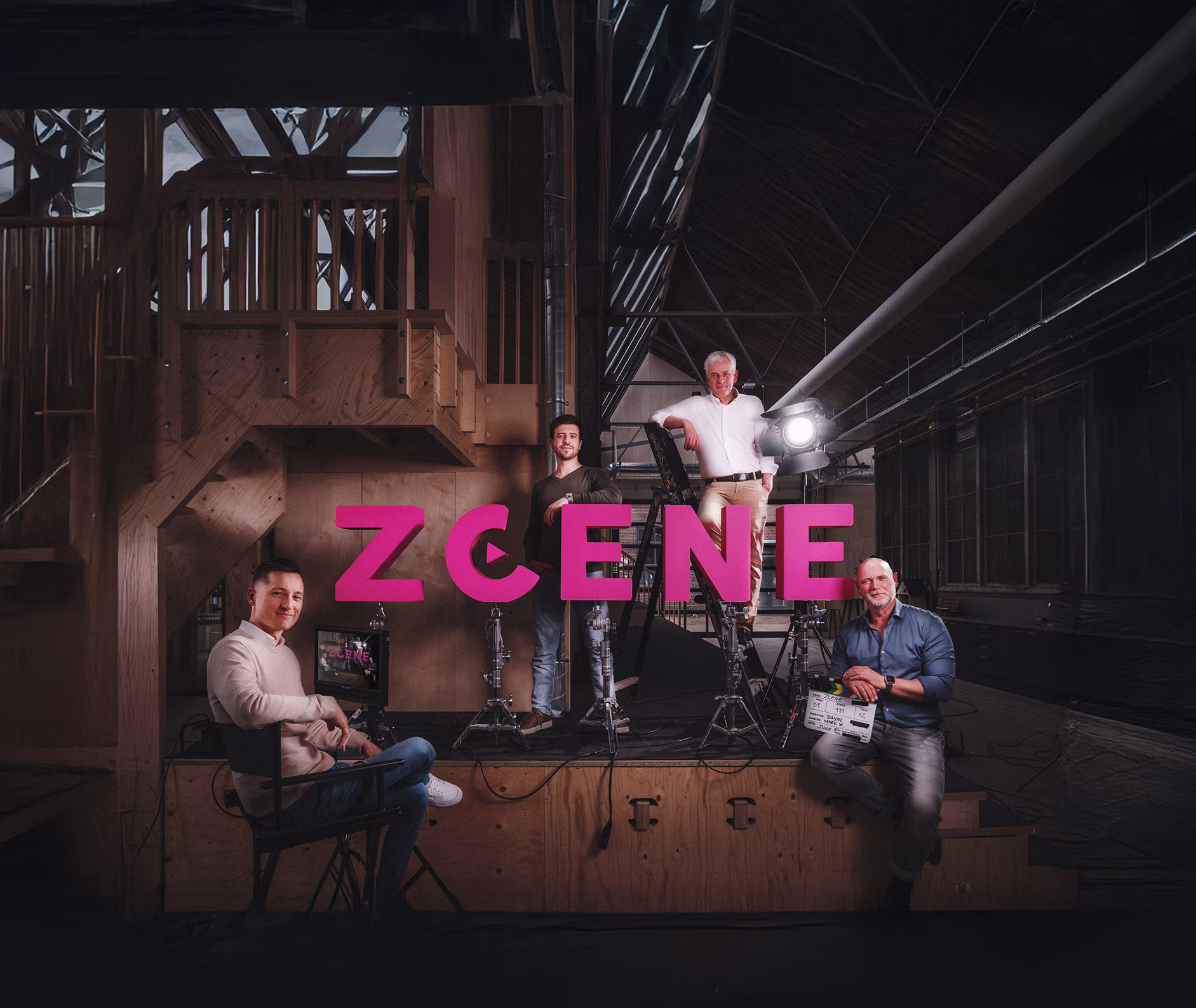 Four men posing on and around a stage with large pink letters spelling ZCENE in an industrial indoor setting with wooden structures and film equipment.