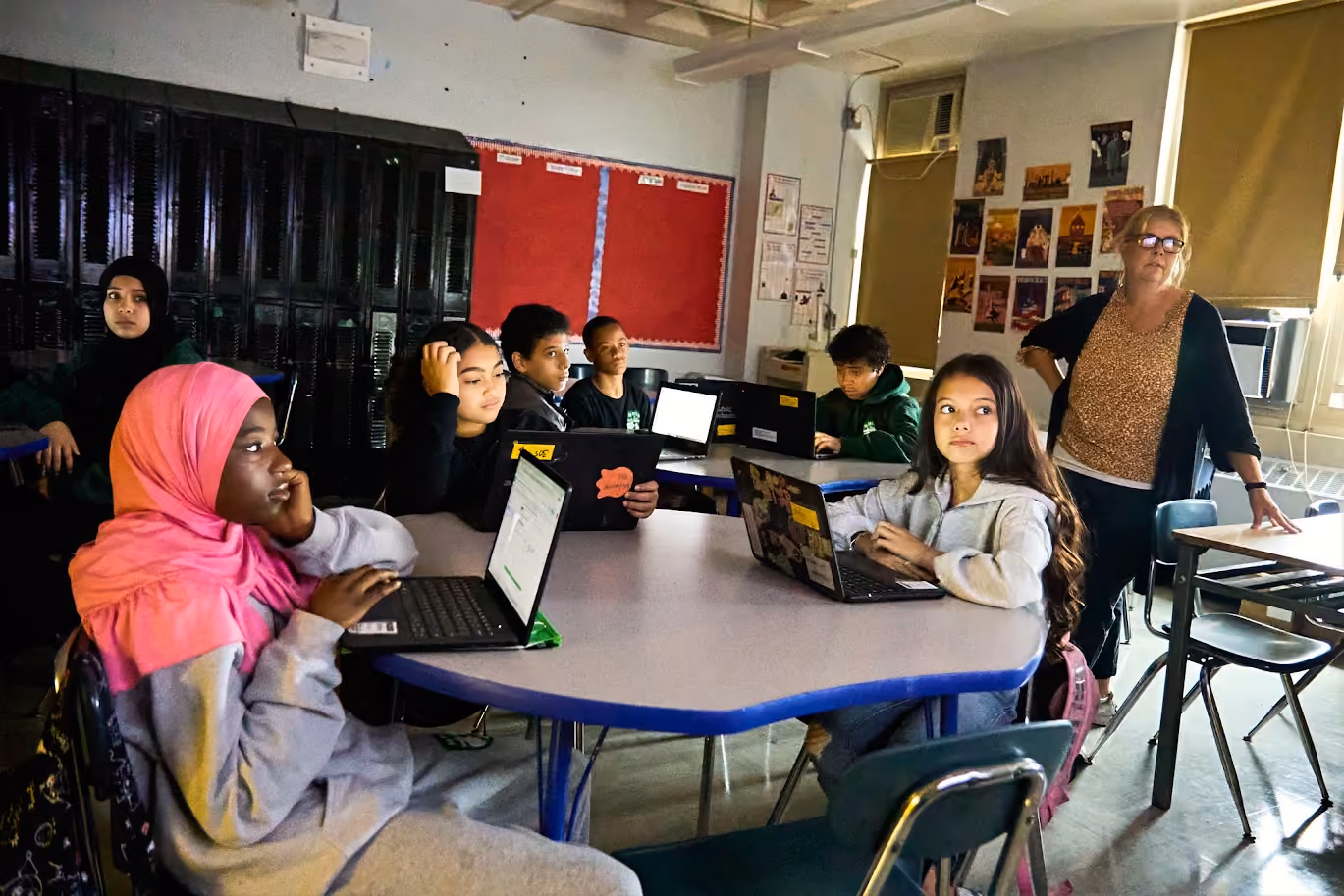 Students work on laptops while teacher observes during classroom lesson