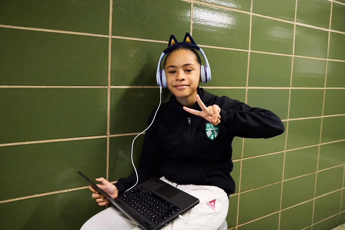 Student wearing headphones holds laptop and flashes peace sign