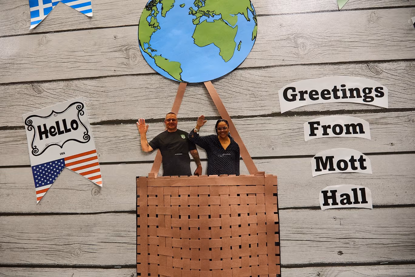 Two staff members waving inside a Greetings From Mott Hall display