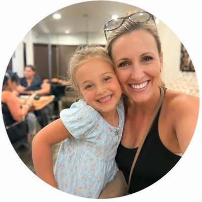 Smiling woman and young girl hugging indoors with people and tables in the background.