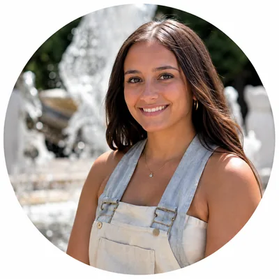 Smiling young woman with long brown hair wearing light overalls standing outdoors near a fountain.