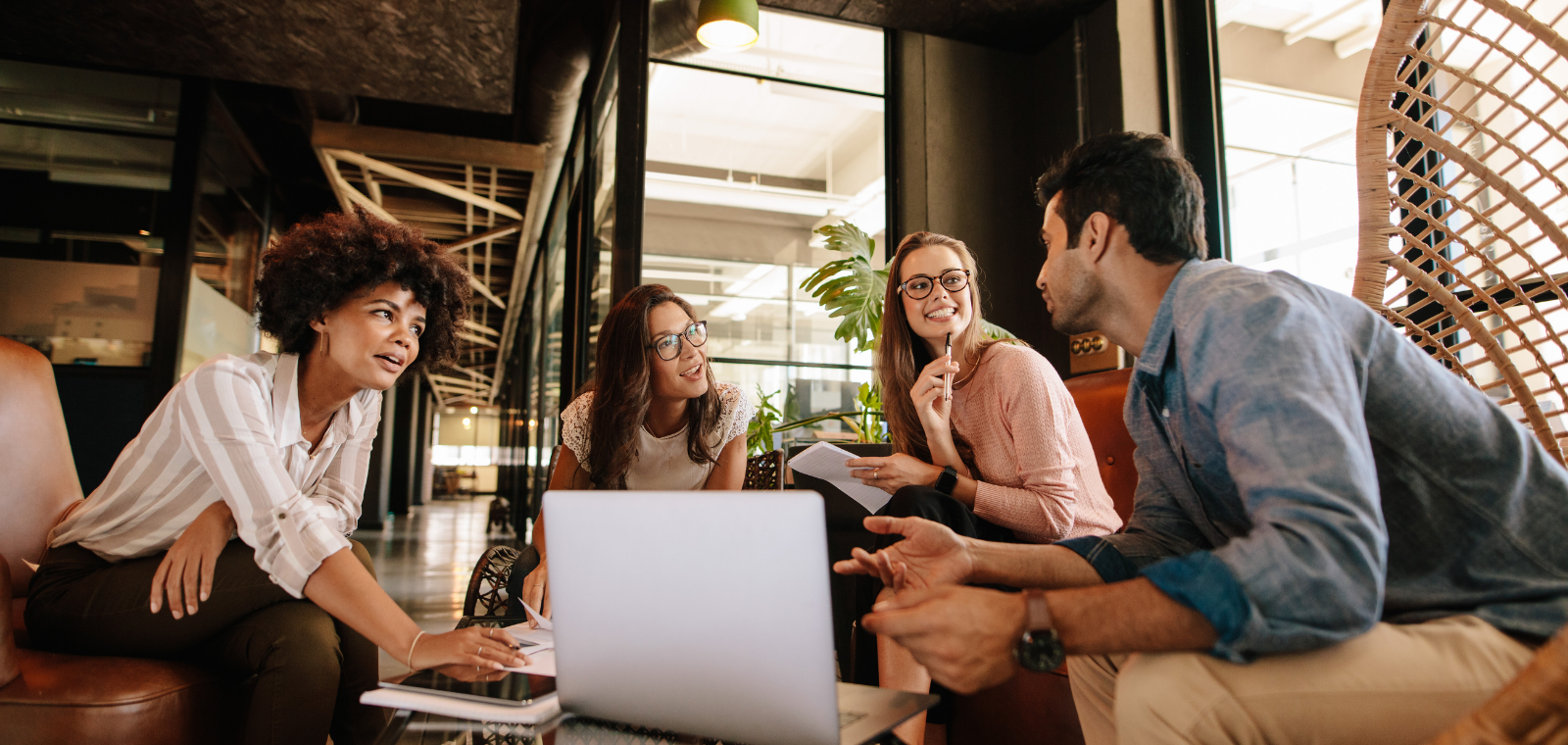 Four diverse young professionals in casual attire engaged in a discussion around a laptop in a modern office lounge.