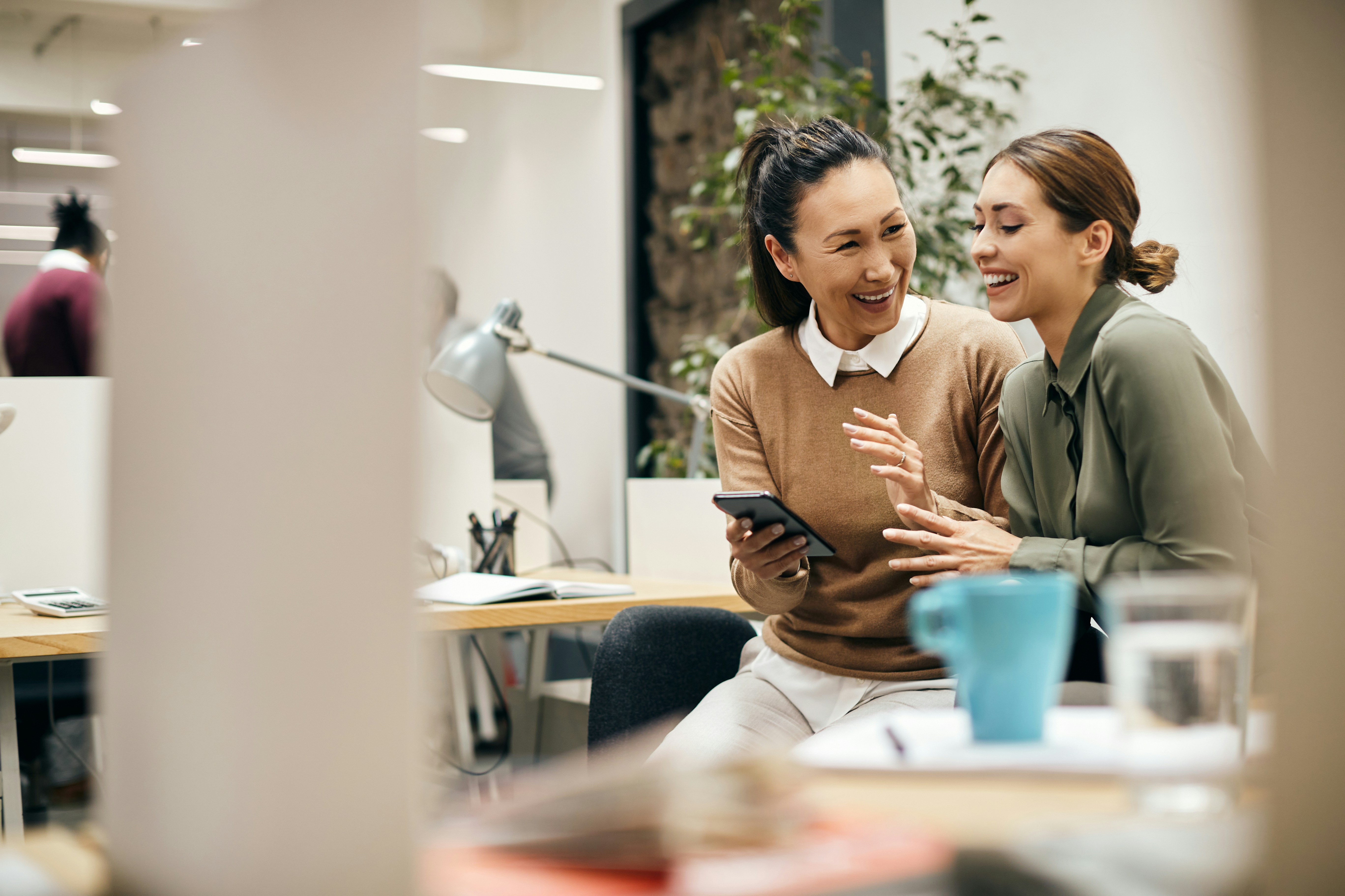 Two women laughing and looking at a smartphone while sitting in a modern office space.