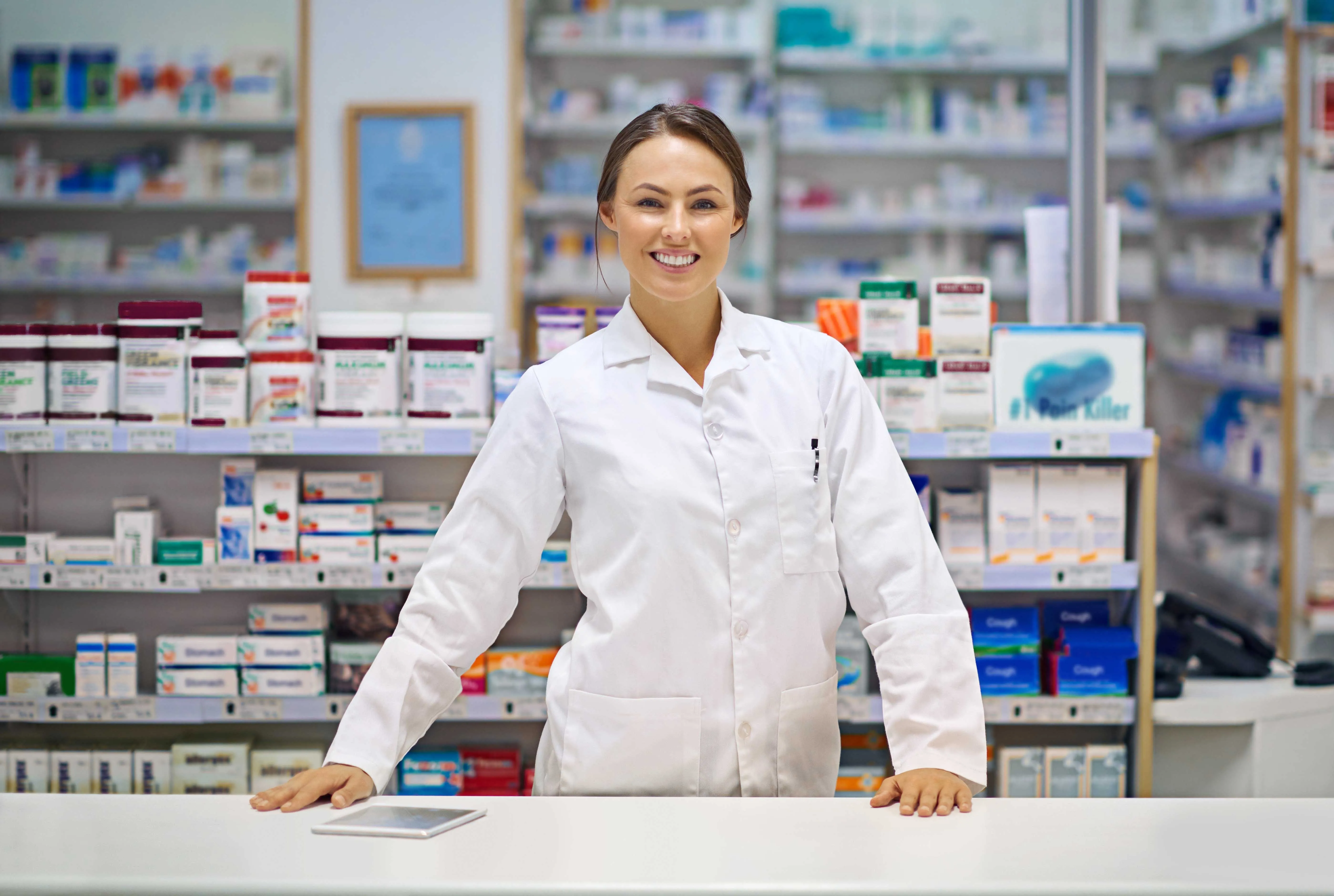 A pharmacist in a white coat standing at a pharmacy counter in front of shelves of stocked medication
