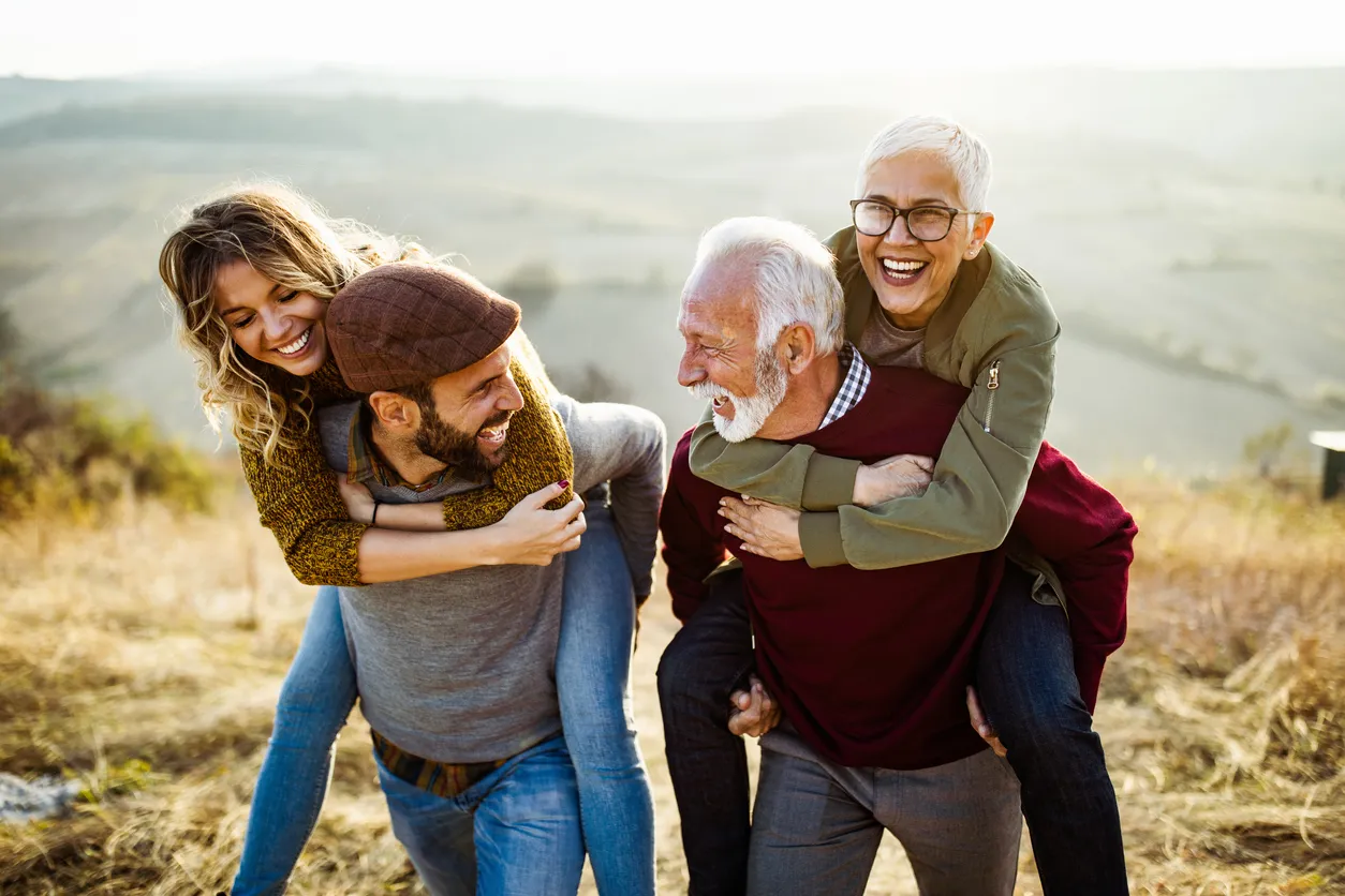 Two couples laughing and giving piggyback rides outdoors on a sunny day