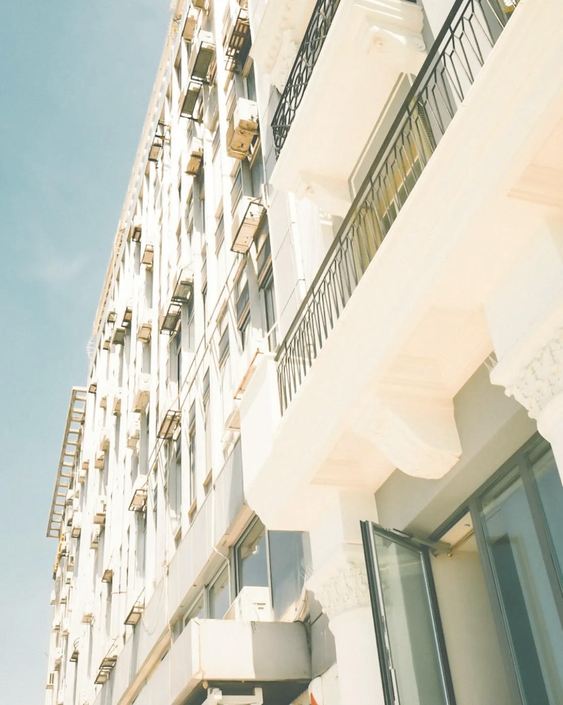Upward view of a white multi-story building with black metal railings and open glass door under clear blue sky.