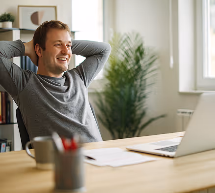 A man is sitting at the table with his hands on the back of his head, in relief, while looking at his computer.