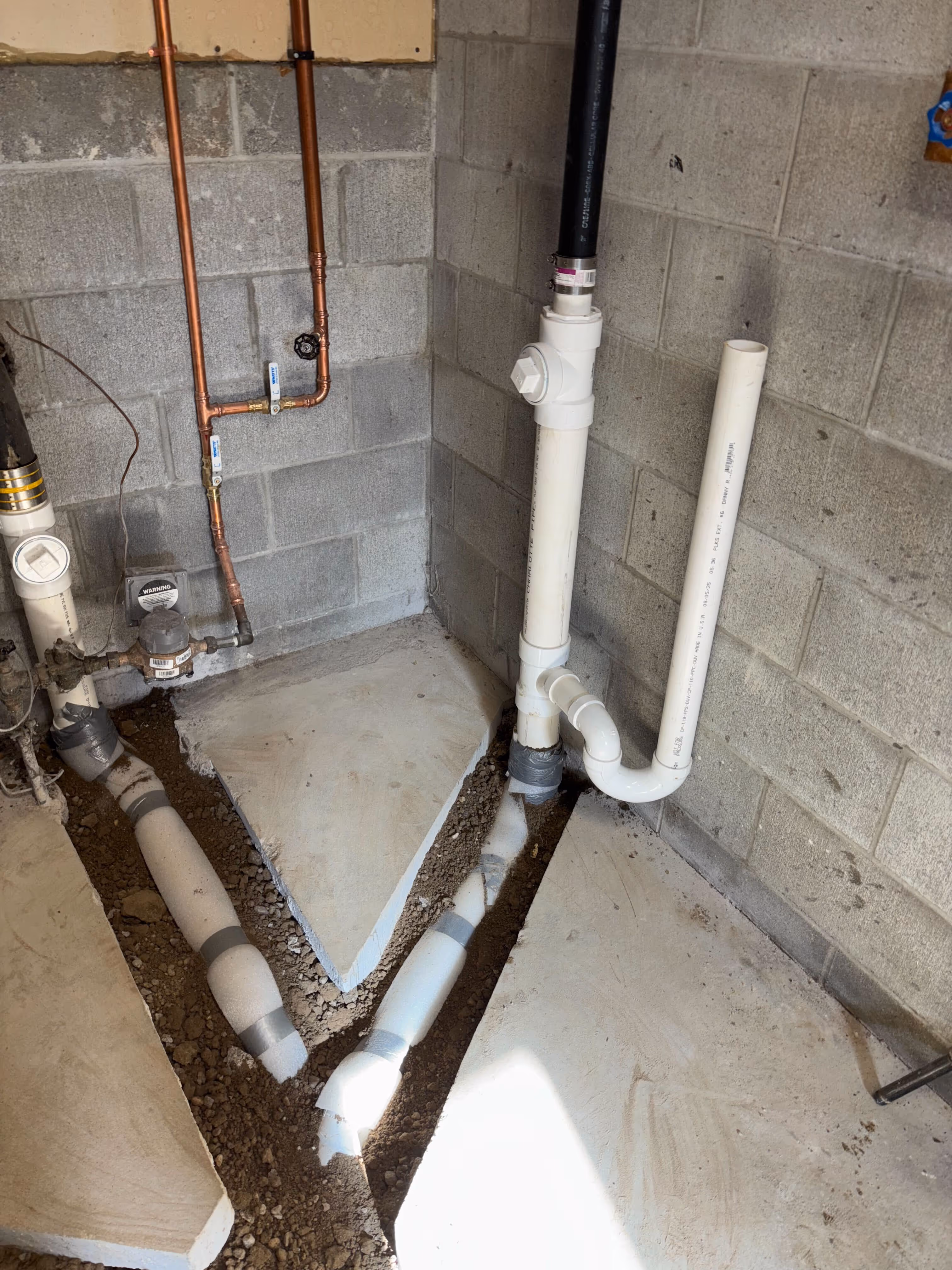 Basement corner where an old black cast iron stack meets new white PVC sewer pipe running into a trench in the concrete floor