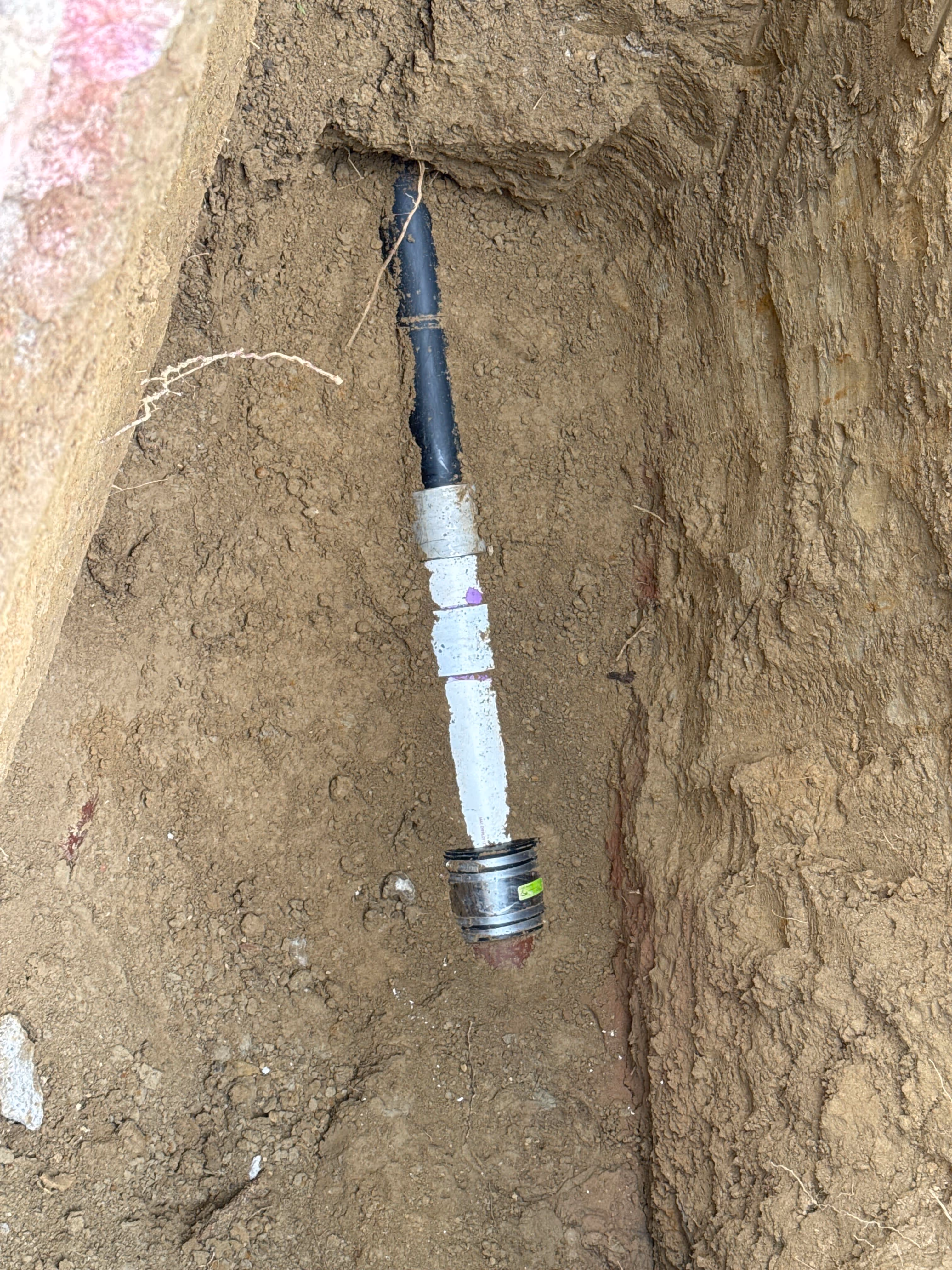 Old black sewer pipe joined to new white PVC pipe with a rubber coupling at the bottom of a dirt trench