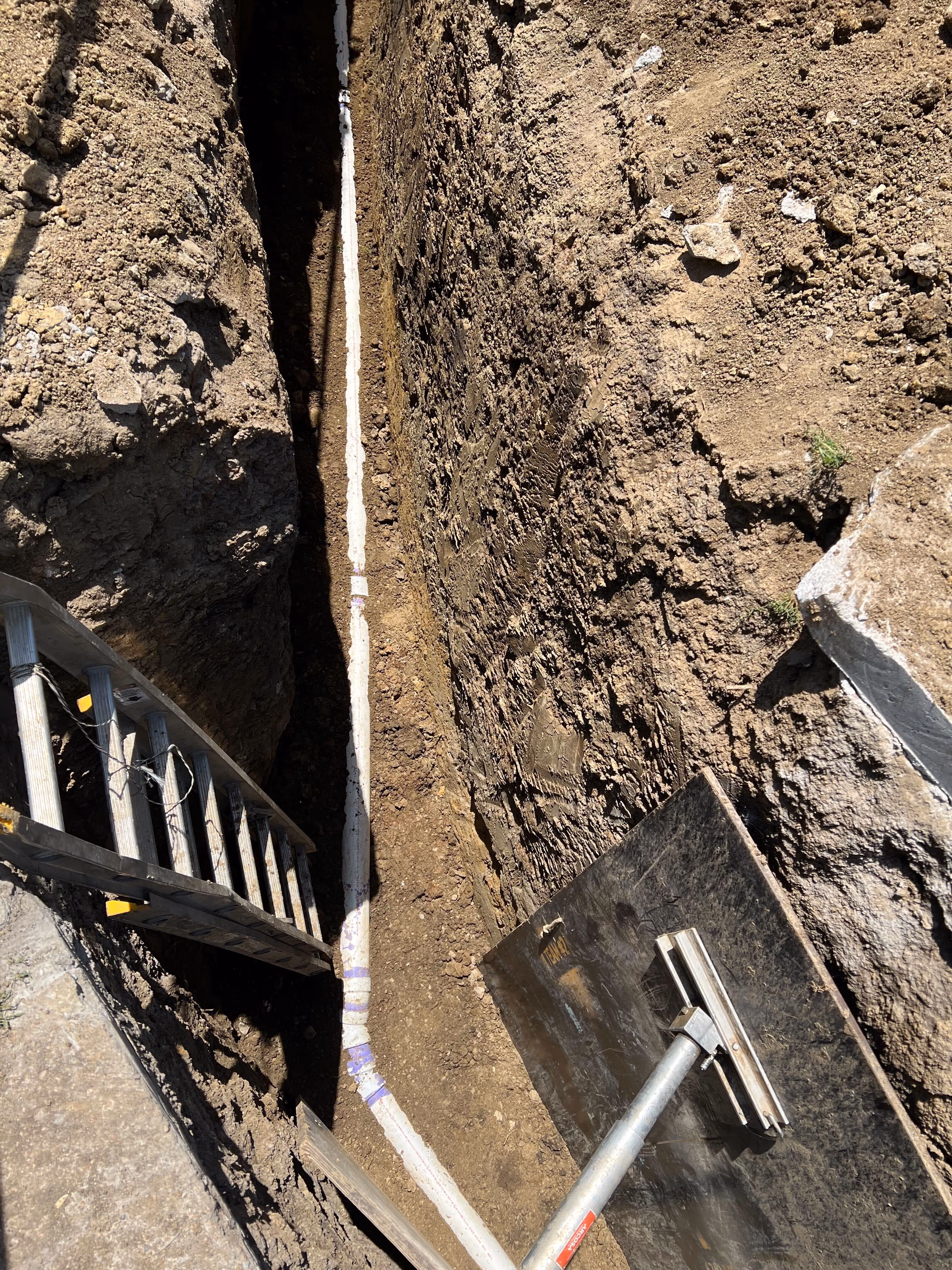 Deep sewer replacement trench with new white sewer pipe laid along the bottom and a ladder leaning on the dirt wall