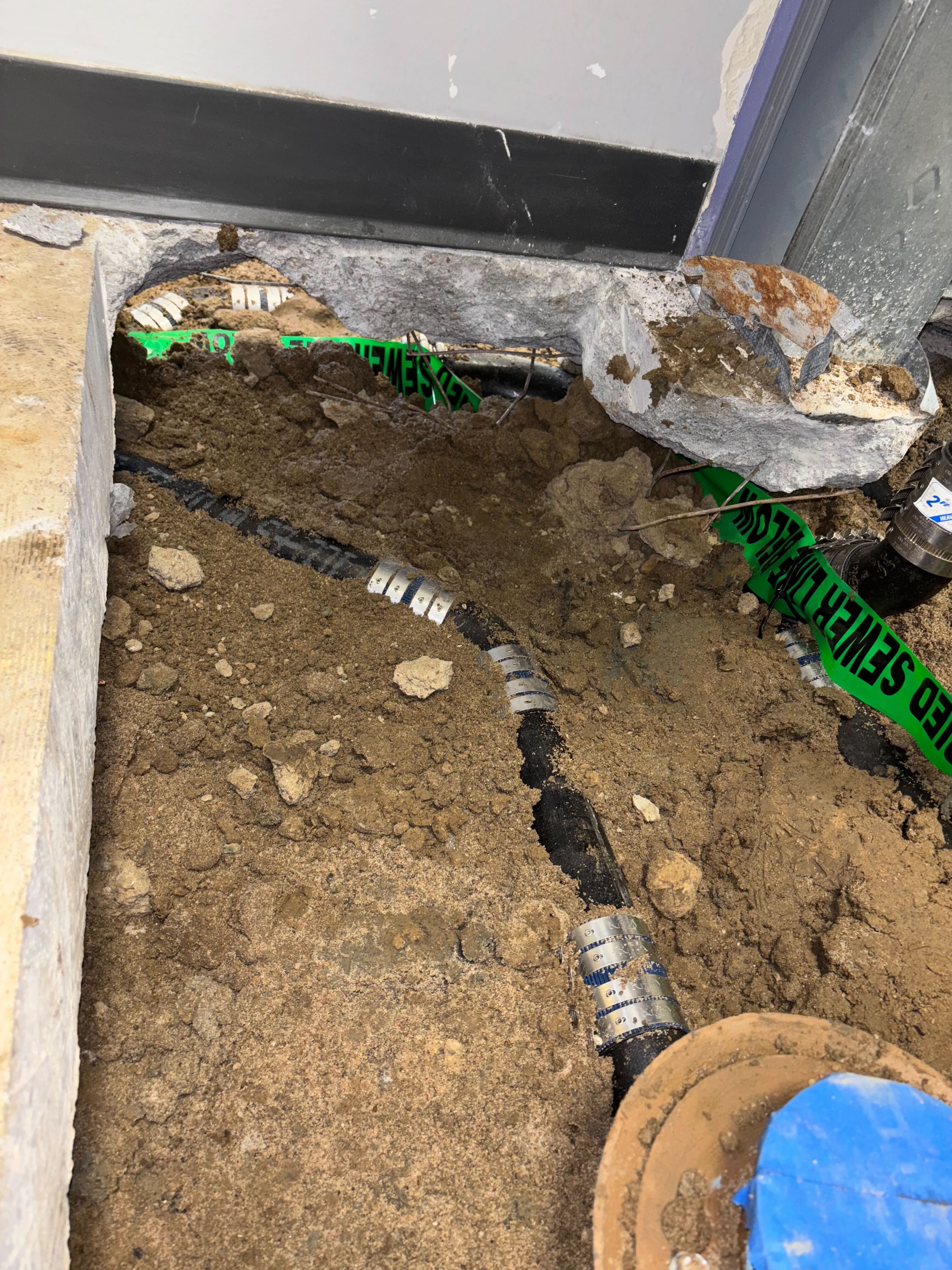 Black cast iron sewer pipe with steel couplings exposed in a dirt trench under a basement floor