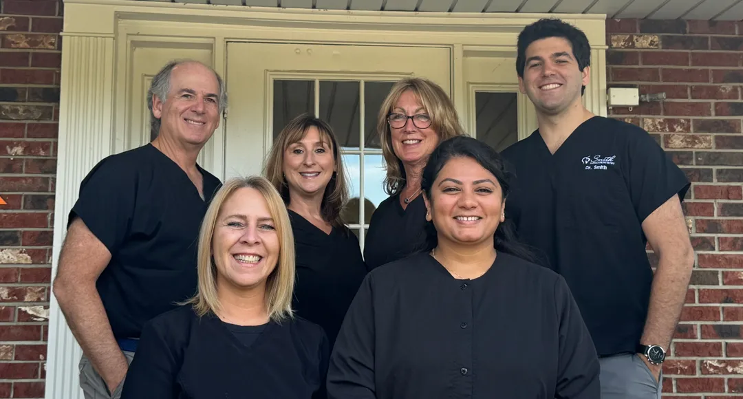 Six smiling medical professionals wearing black scrubs standing in front of a door with glass panels and brick walls.