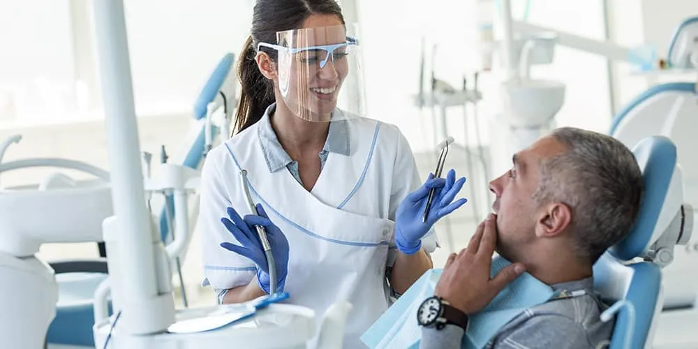 Dentist wearing gloves and face shield talking to a male patient seated in a dental chair.