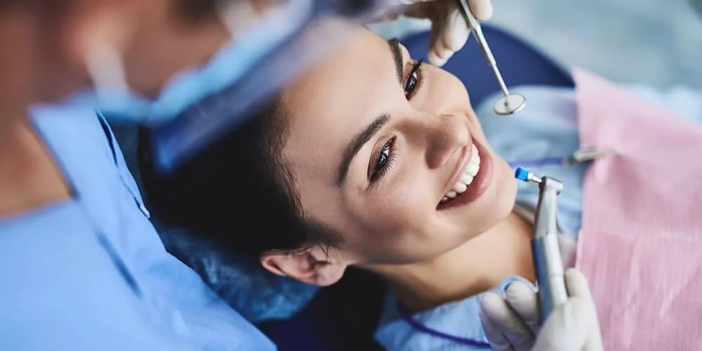 Smiling woman lying back in dental chair receiving dental checkup from dentist wearing gloves.