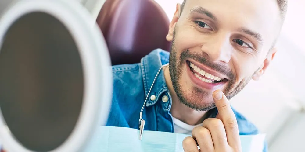 Smiling man pointing to his teeth while sitting in a dental chair looking into a handheld mirror.