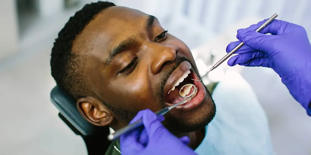 Dentist with purple gloves examining the open mouth of a male patient using dental mirror and probe.