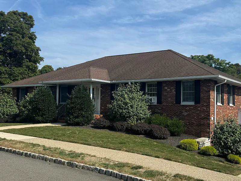 Single-story brick house with dark shutters, a brown roof, and landscaped front yard with bushes and a sidewalk.
