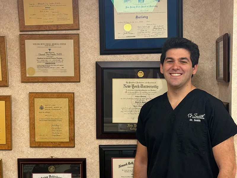 Smiling male dentist in black scrubs stands in front of a wall displaying multiple framed dental degrees and certificates.
