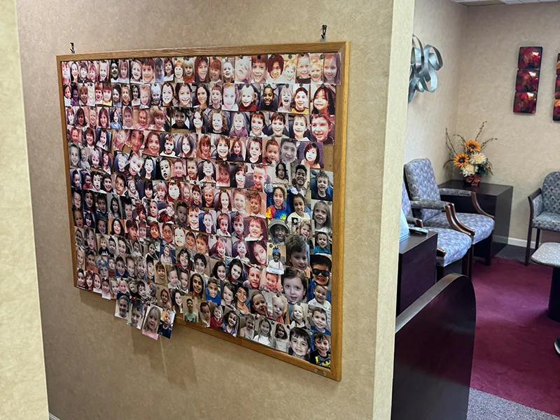 A bulletin board mounted on a beige wall covered with numerous small photographs of smiling children and some adults in a waiting room with upholstered chairs and decorative wall art.