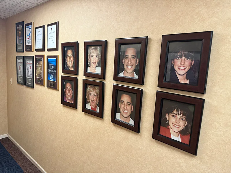 Wall with framed awards, certificates, and portraits of smiling people in an office hallway.