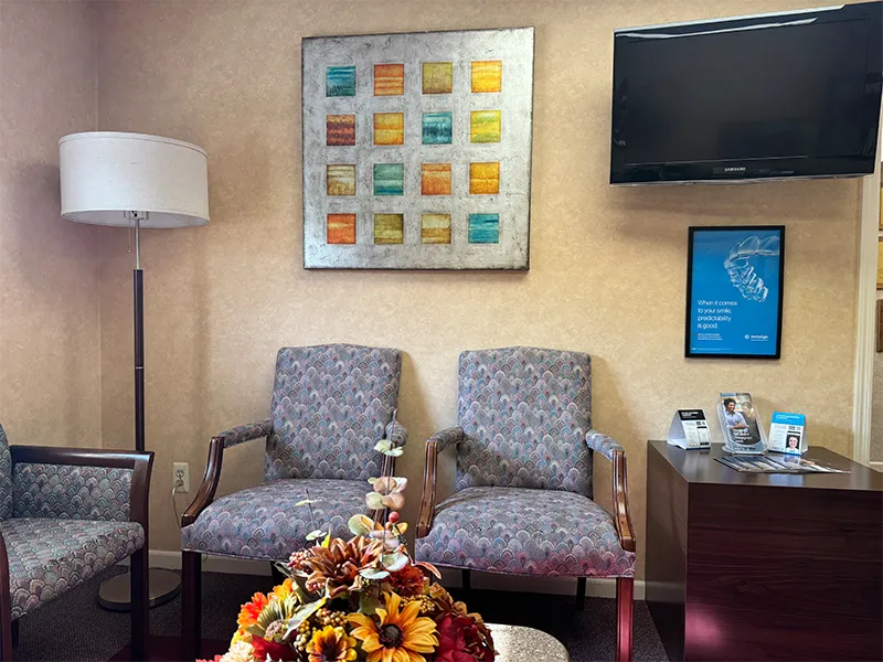 Waiting room with three patterned chairs, a floor lamp, a colorful flower arrangement on a table, a mounted TV, and informational brochures on a wooden side table.