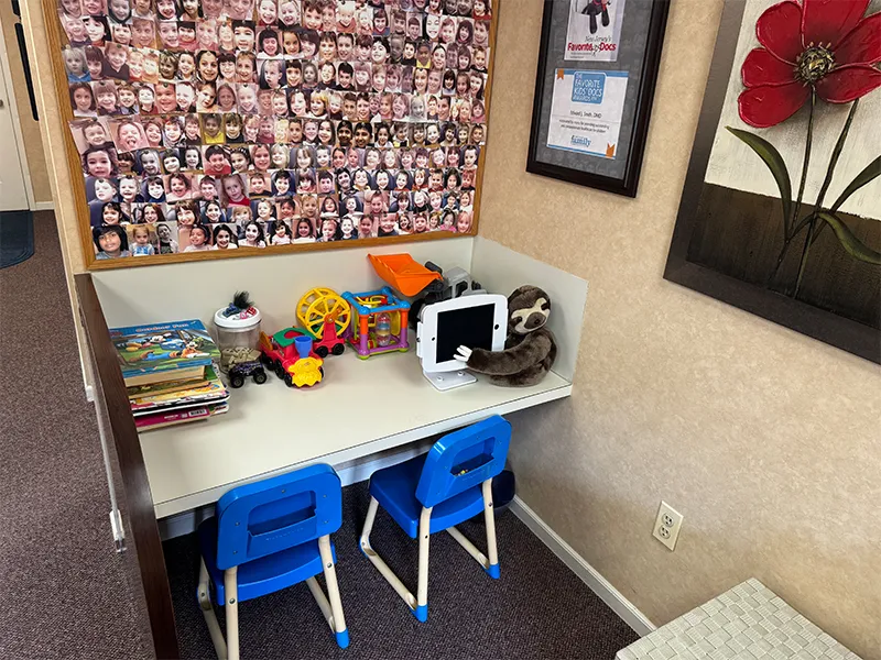 Children's play corner with two blue chairs, toys on a white table, a stuffed sloth hugging a toy tablet, and a wall covered in many children's photos.