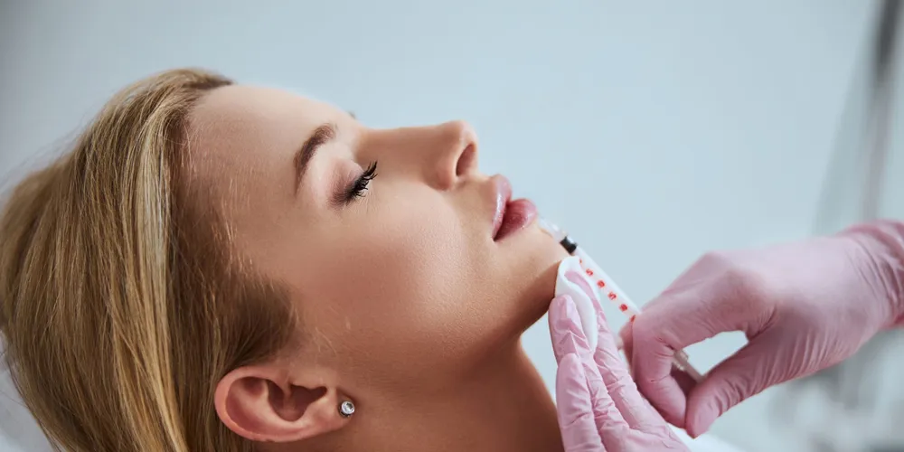 Close-up of a woman receiving a cosmetic injectable treatment on her chin by a practitioner wearing pink gloves.