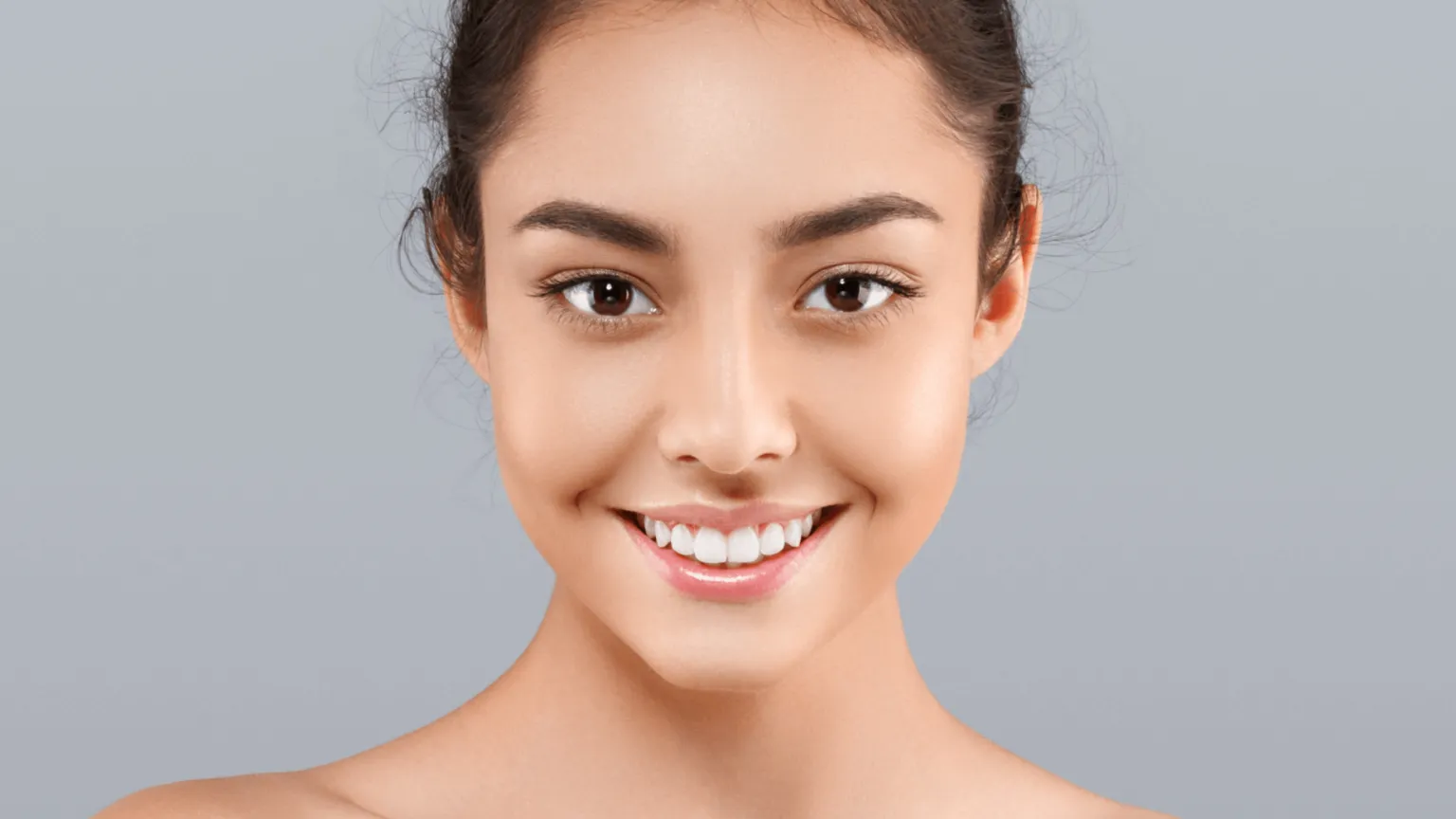 Close-up portrait of a young woman with clear skin and a bright smile against a gray background.