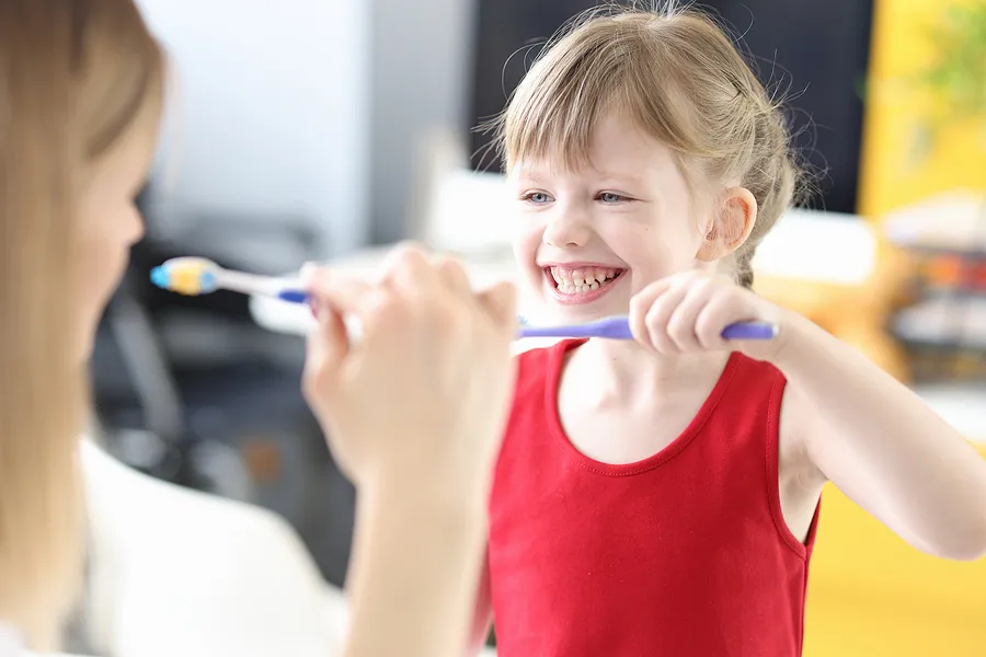 Smiling young girl in a red sleeveless shirt holding a toothbrush with toothpaste, facing an adult woman.