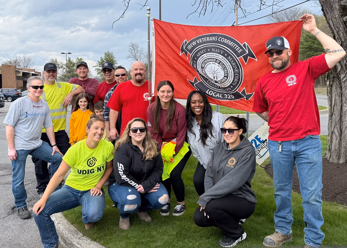 A diverse group of individuals poses in front of a red flag representing the IBEW Veterans Committee, showcasing camaraderie and community spirit.