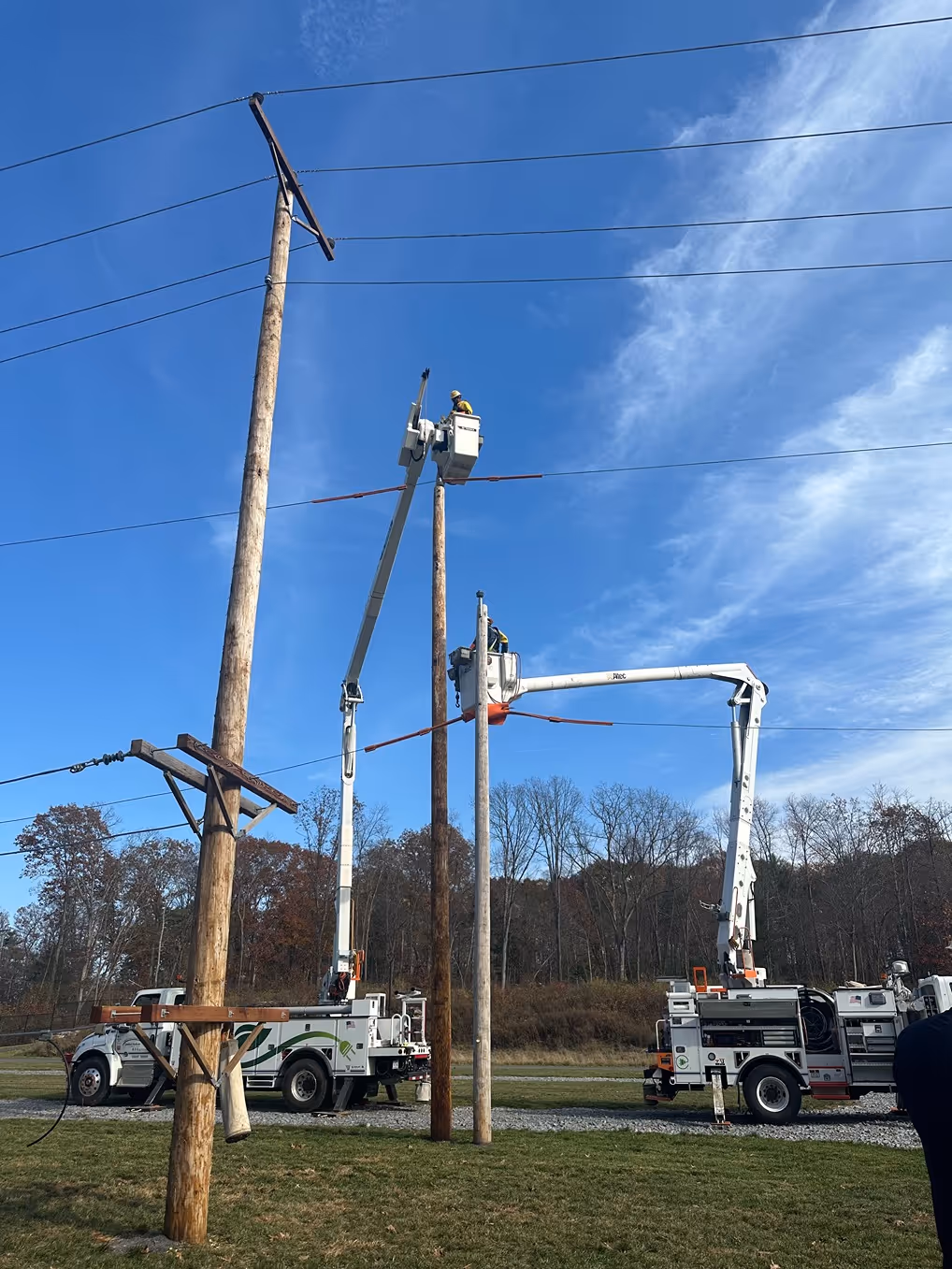 Two utility trucks with aerial lifts working on power lines between wooden poles under a clear blue sky.