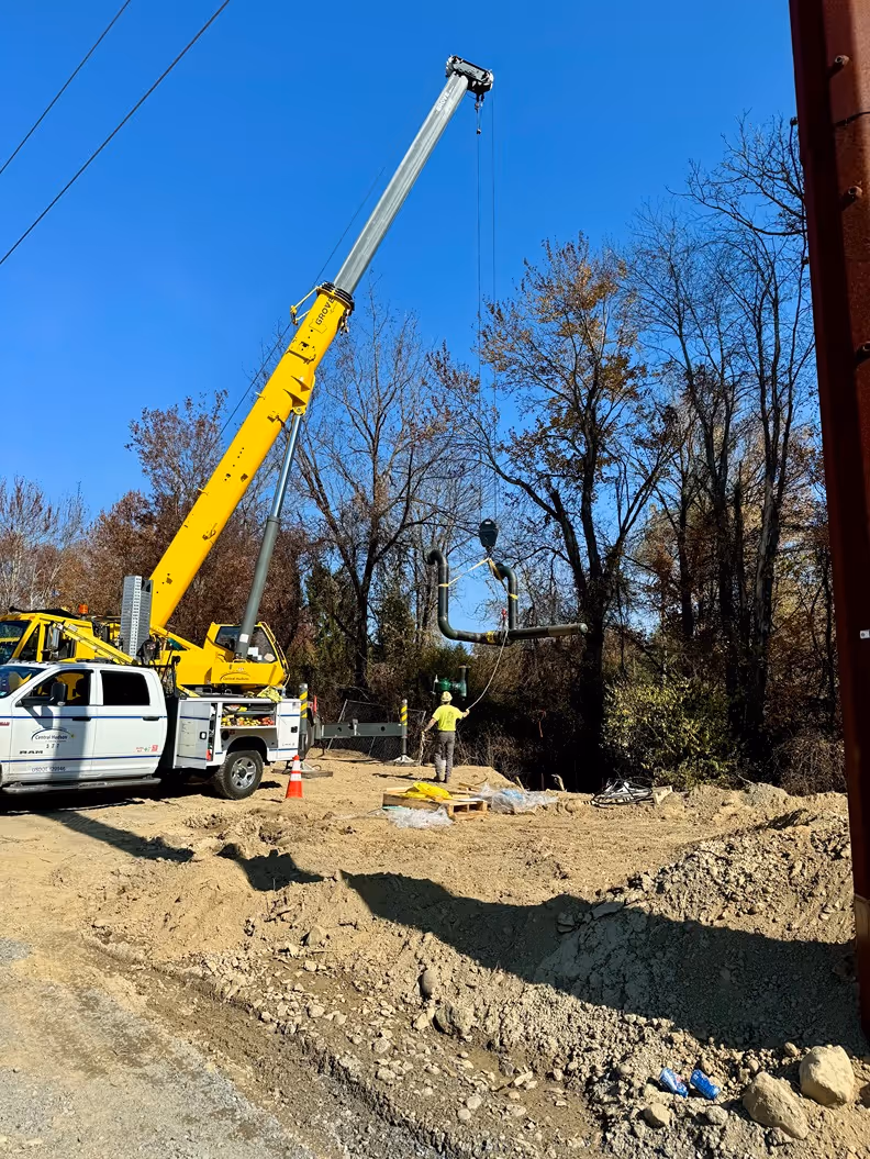 A yellow crane lifts materials at a construction site, while a worker in a yellow shirt oversees the operation under a clear blue sky.