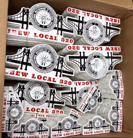 A box filled with white stickers featuring the IBEW Local 320 logo and a bridge design, stacked neatly on top of each other.