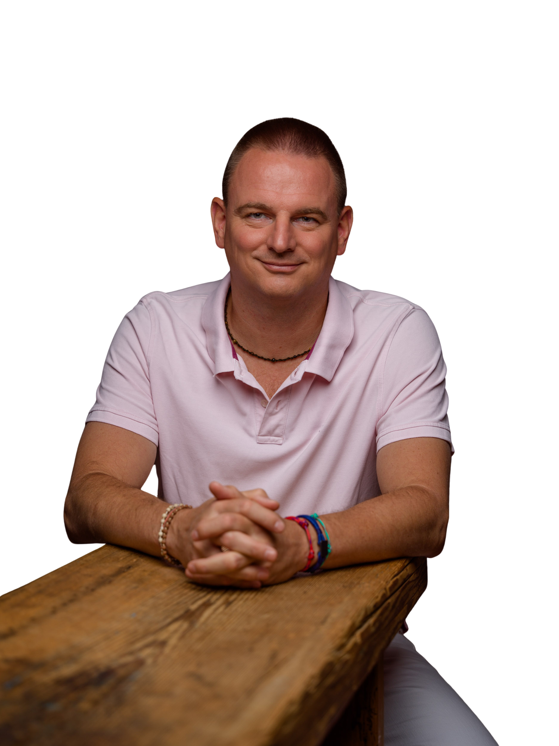 Mann mit rosa Poloshirt und bunten Armbändern sitzt lächelnd an einem rustikalen Holztisch.