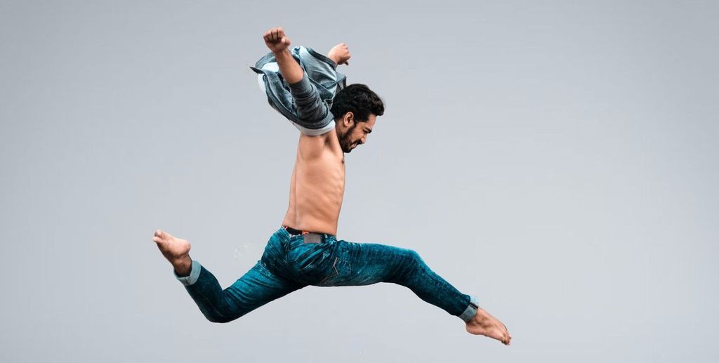 Ein muskulöser Mann im Sprung, der sein Hemd auszieht, vor einem grauen Hintergrund.