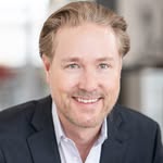 Smiling middle-aged man with short gray hair wearing a gray checkered suit jacket and a light blue checked shirt.
