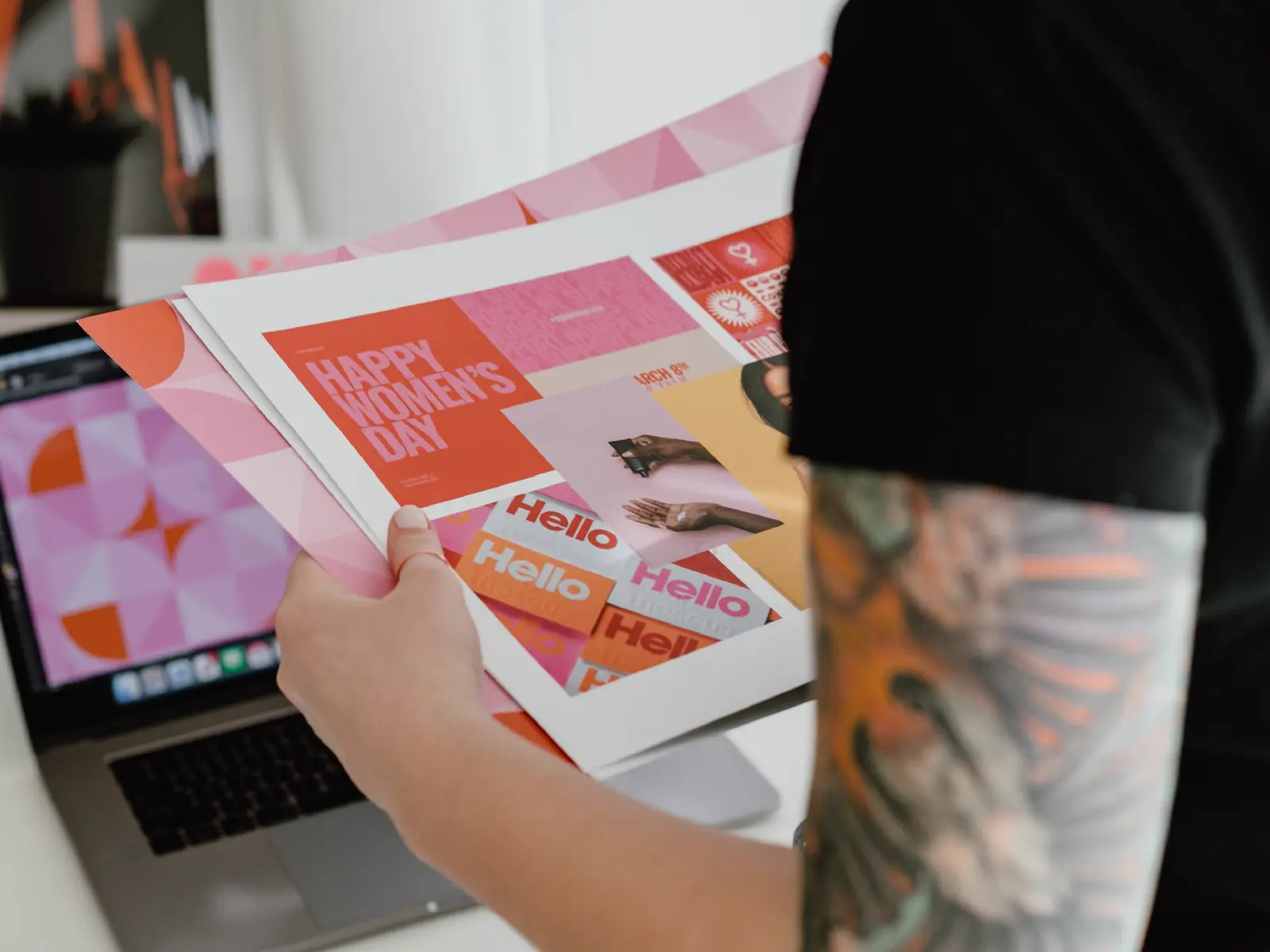 Person holding colorful printed graphic designs near a laptop displaying a pink geometric pattern.