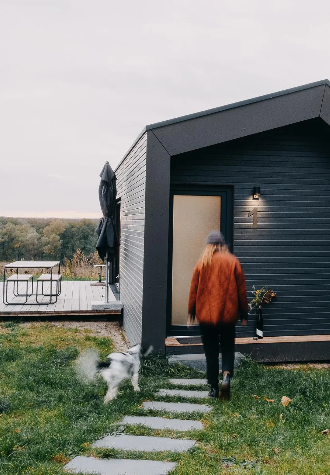 Person walking toward a dark modern cabin while a dog runs beside them on the grass.