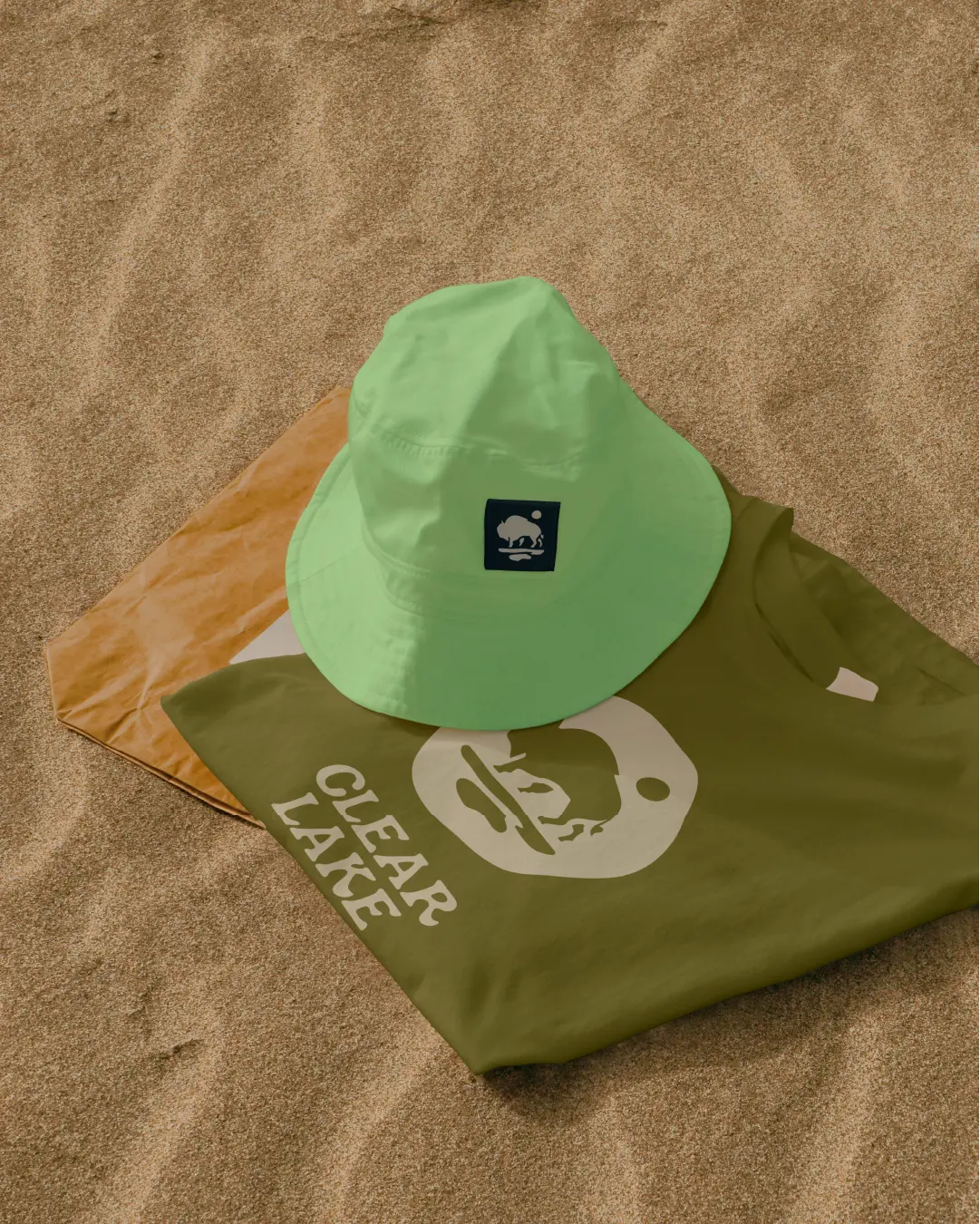 Close-up of Clear Lake Country branded bucket hat and t-shirt on sandy beach, featuring the bison mark.