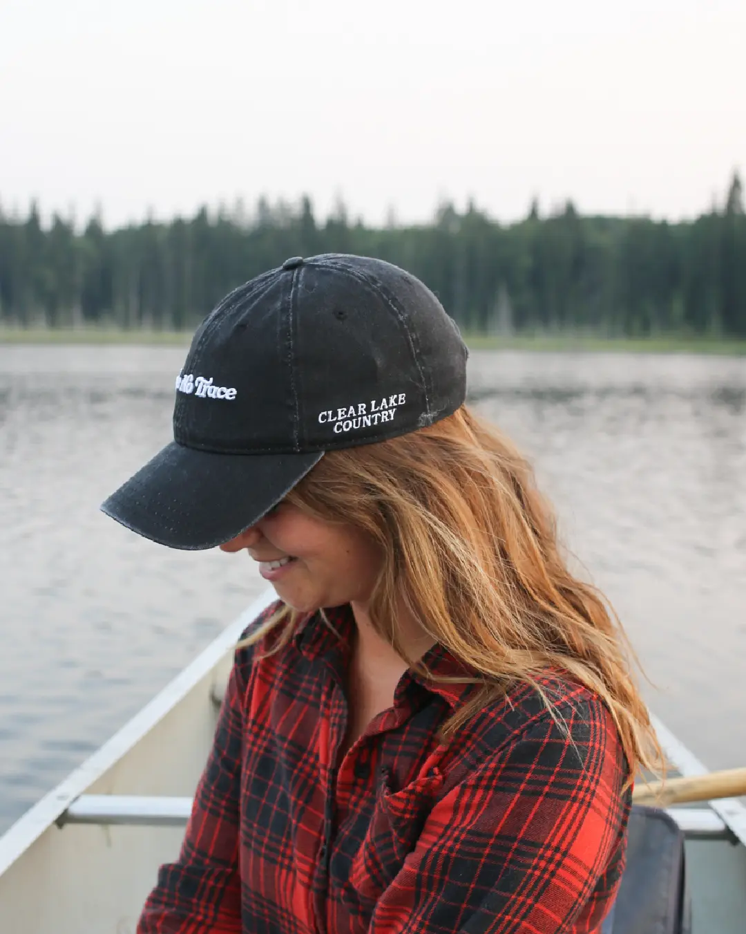 Person sitting in a canoe wearing a black cap embroidered “Clear Lake Country,” with lake and forest in the background.