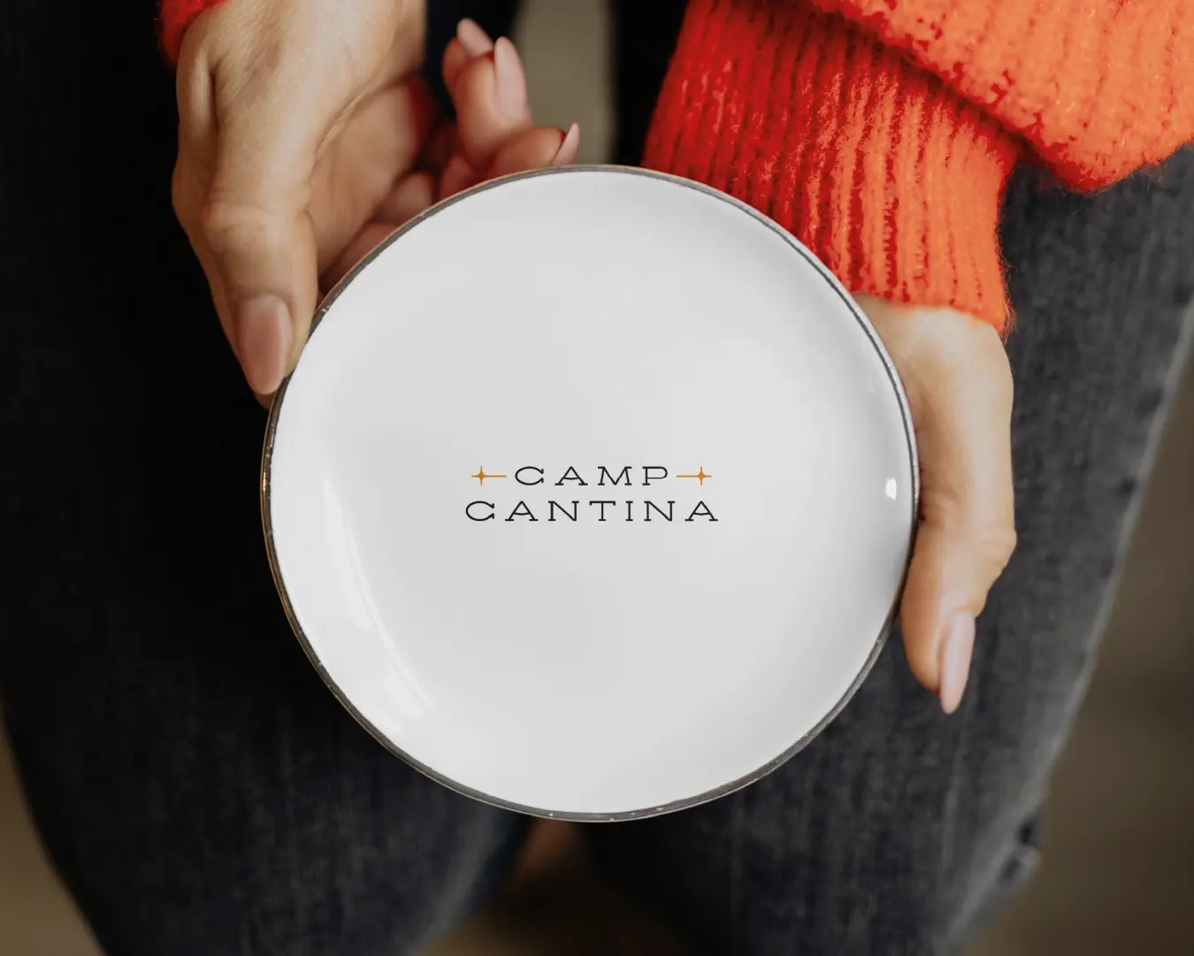 Hands holding a white plate featuring the Camp Cantina wordmark with small star-like icons.