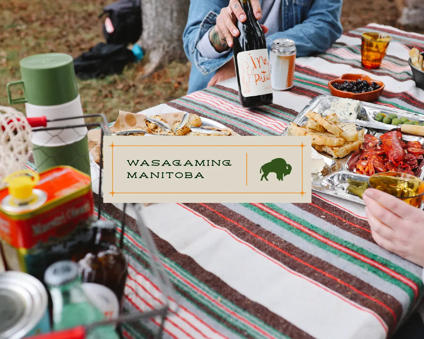 Picnic table spread with wine and snacks, featuring a “Wasagaming, Manitoba” label and bison icon.