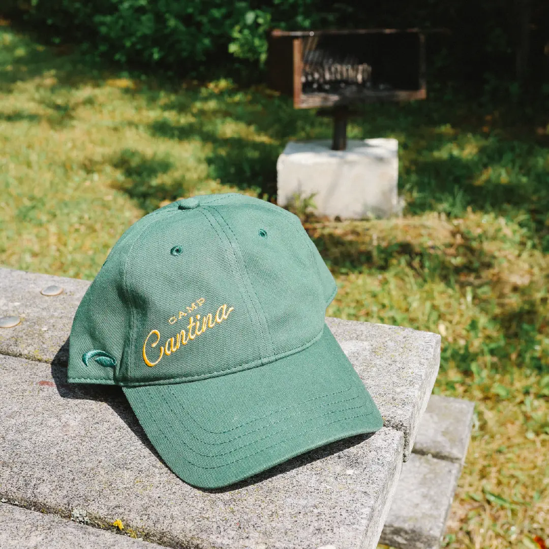Green Camp Cantina hat with orange embroidered script logo, photographed outdoors.