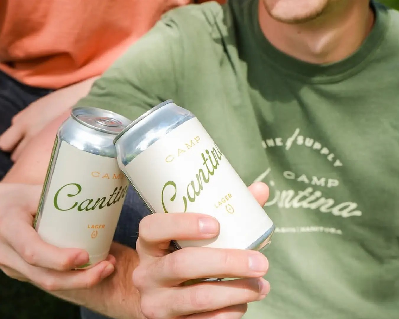 Two people clinking Camp Cantina Lager cans, showing the Camp Cantina script label up close.