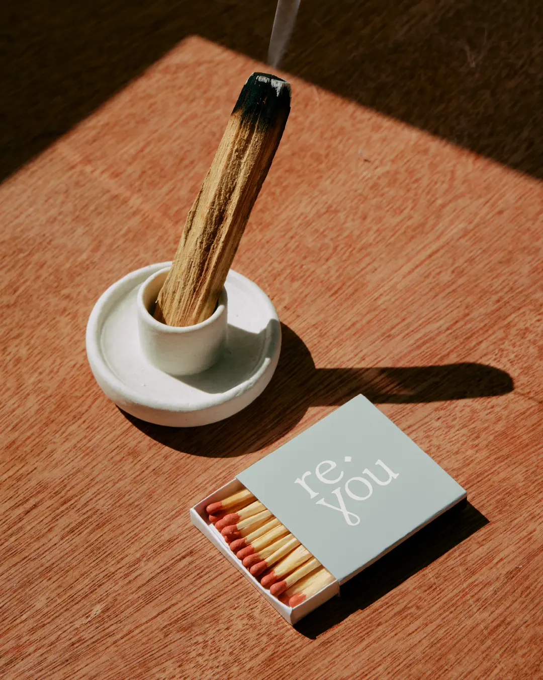 Smouldering wooden incense stick in a ceramic dish beside a Reyou-branded matchbox on a warm wood tabletop.