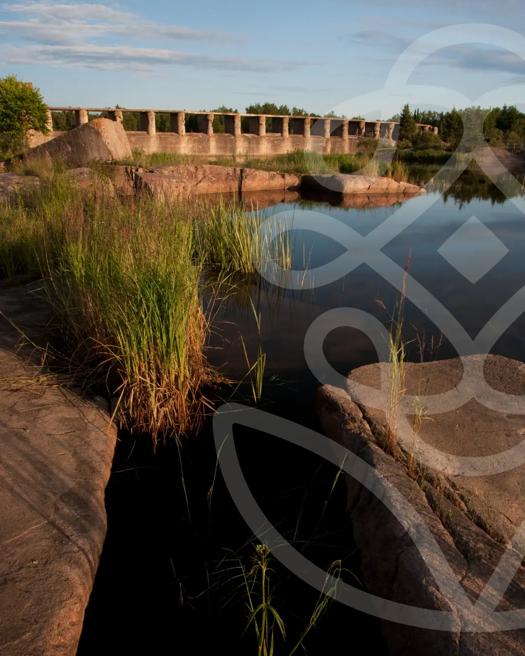 Calm water framed by rock and grasses with a historic stone bridge in the background in Pinawa, Manitoba, overlaid with a subtle Reyou icon.