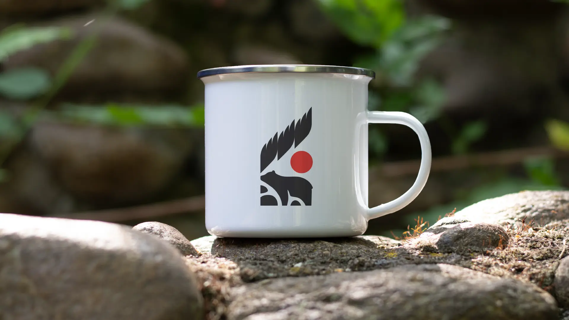 White enamel camp mug featuring the Mino Kanawapamin logo, resting on rocks outdoors.
