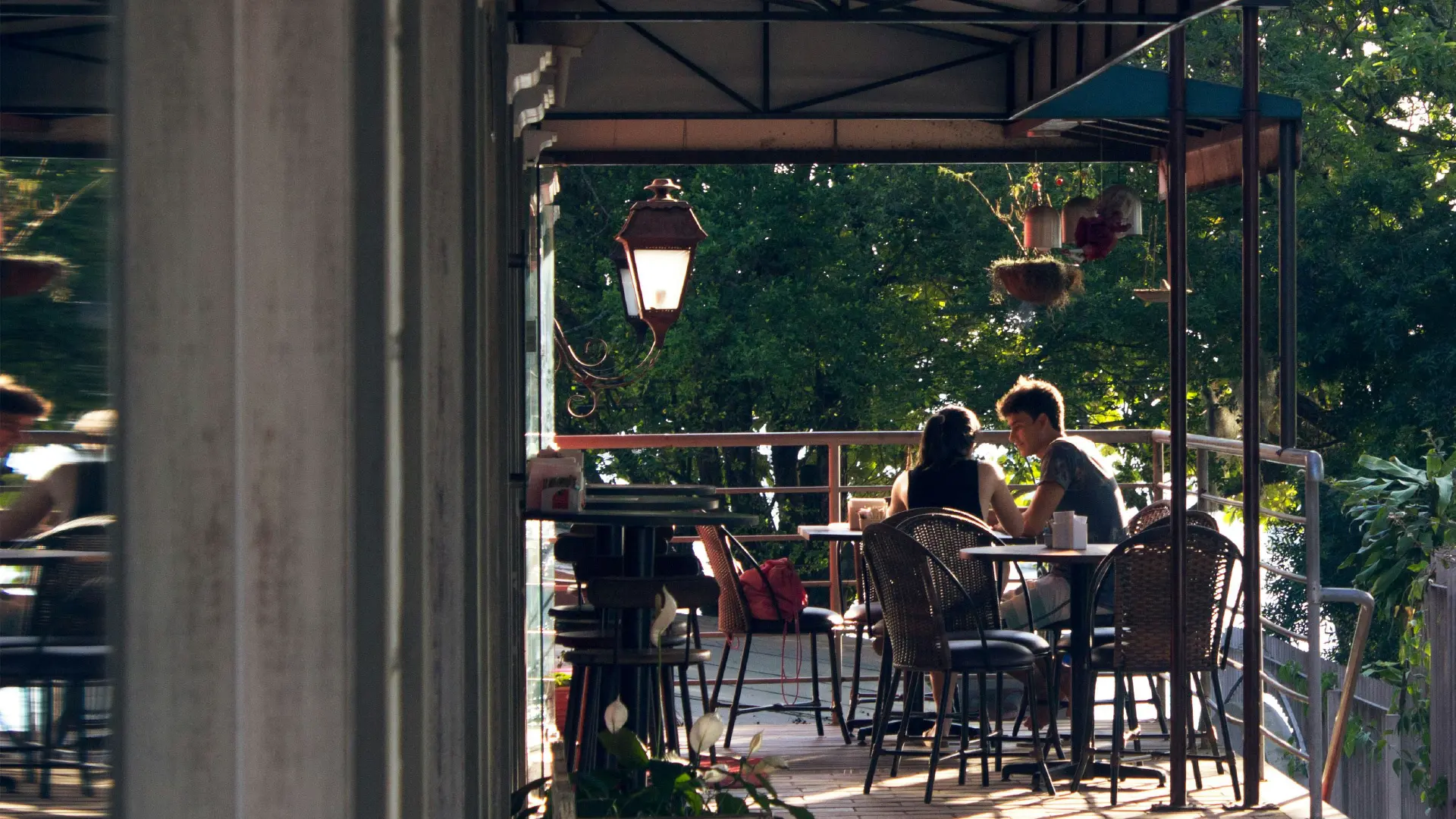 Two people seated at a restaurant patio table under a covered terrace, surrounded by greenery and warm afternoon light.